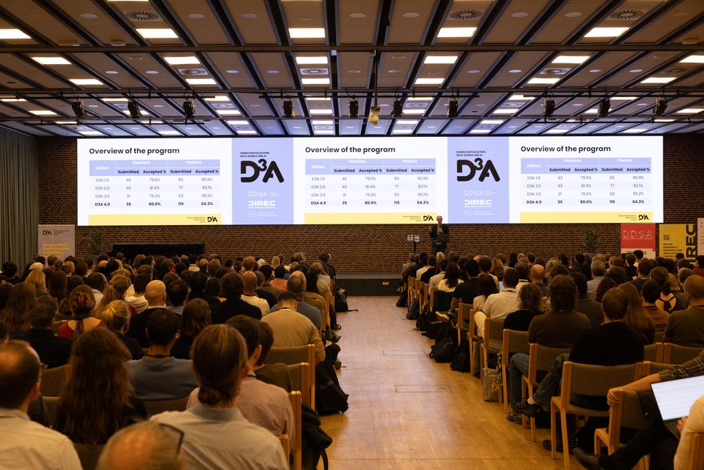 Audience attending a D3A conference in a large auditorium while a presenter shares program statistics on three large screens.